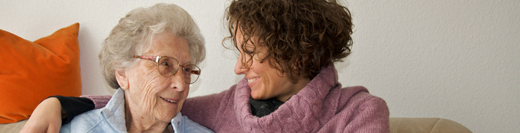 Fürsorgliches Treffen von zwei Frauen aus unterschiedlichen Generationen - sitzen auf einem Sofa - die jüngere Frau umarmt die ältere Frau